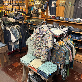 A cluttered clothing store displays a variety of patterned shirts, neatly folded pants, and caps. The atmosphere suggests an outdoor or adventure theme.