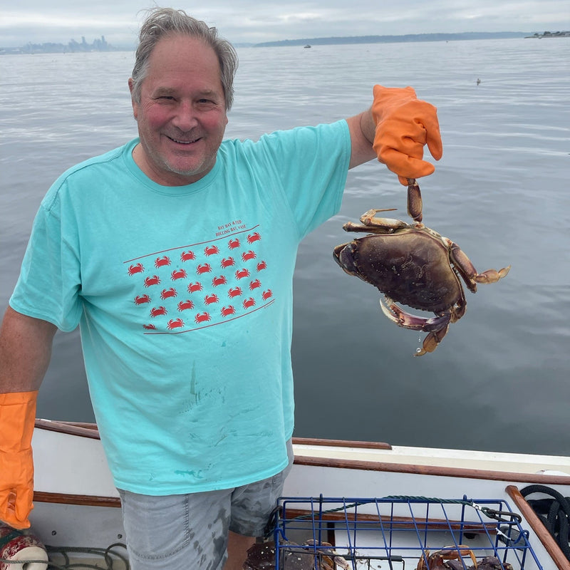 A smiling man in a turquoise crab-patterned shirt and orange gloves holds a large crab on a boat. A crab trap is visible, with calm sea and cloudy sky.