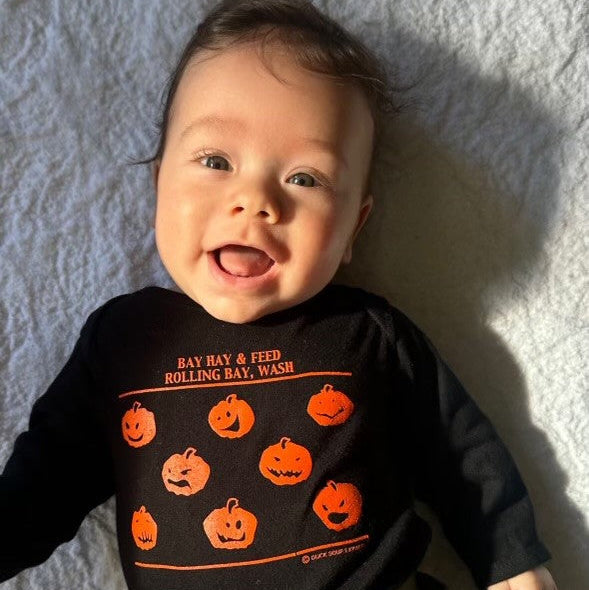 A joyful baby lies on a white blanket, wearing a black shirt with orange pumpkins and text. They are smiling warmly, with bright sunlight illuminating their face.