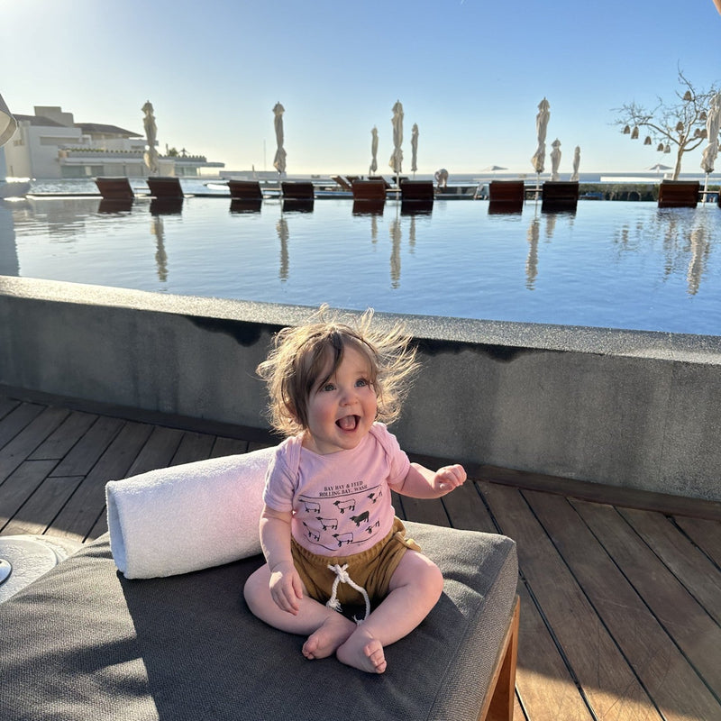 Baby sitting on a lounge chair by an infinity pool, smiling joyfully. Bright blue sky and closed umbrellas in the background evoke a warm, sunny day.
